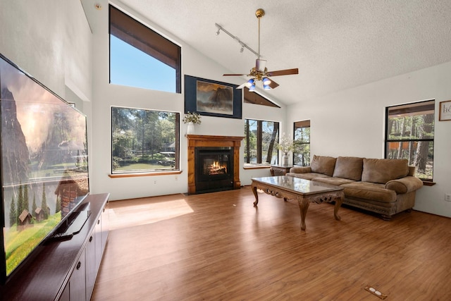 living room featuring high vaulted ceiling, a textured ceiling, wood finished floors, a ceiling fan, and a glass covered fireplace
