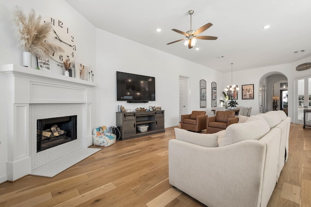 living area featuring light wood-style flooring, visible vents, a tiled fireplace, arched walkways, and ceiling fan with notable chandelier