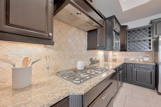 kitchen featuring backsplash, light tile patterned flooring, under cabinet range hood, light stone countertops, and stainless steel gas stovetop