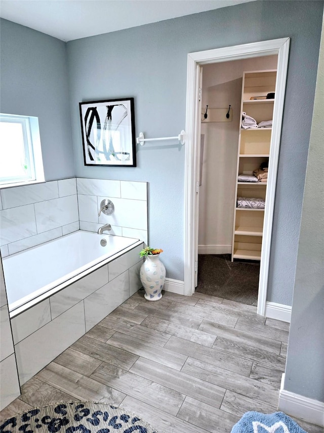 full bathroom featuring wood finished floors, a walk in closet, a garden tub, and baseboards