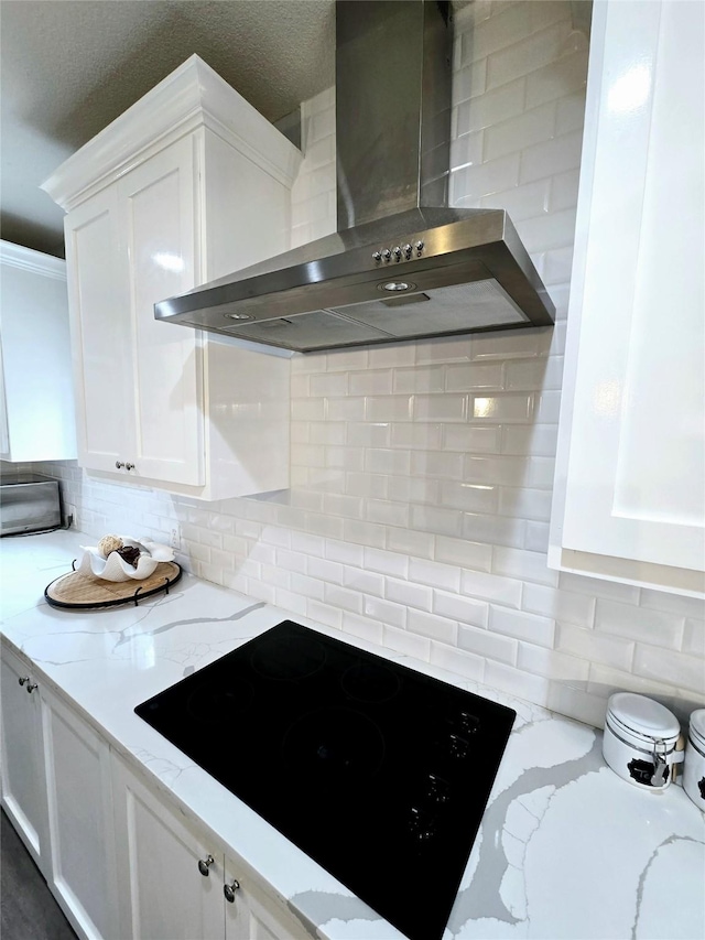 kitchen featuring wall chimney exhaust hood, black electric stovetop, white cabinetry, backsplash, and light stone counters