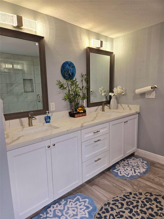 full bathroom featuring wood finished floors, a shower stall, double vanity, baseboards, and a textured wall