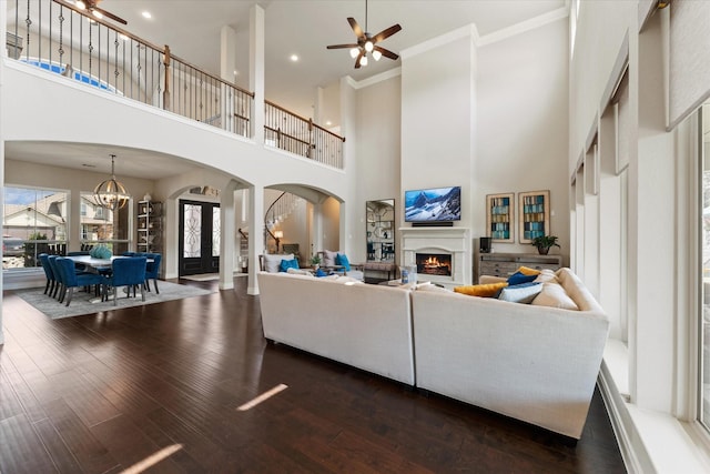 living area featuring dark wood-style flooring, a warm lit fireplace, arched walkways, a high ceiling, and ornamental molding