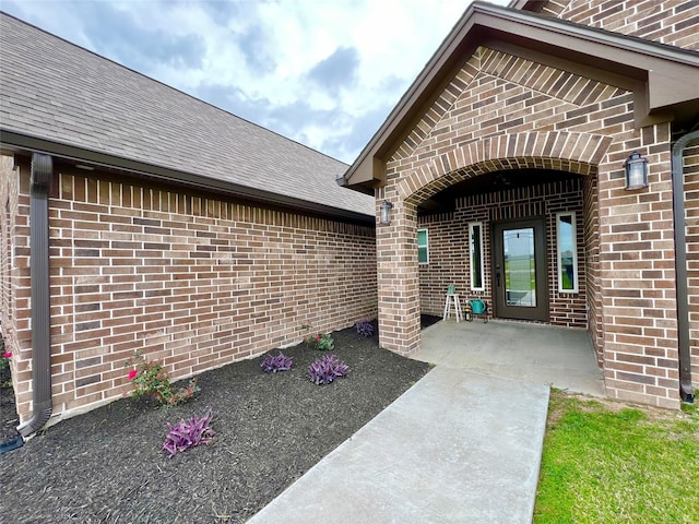 entrance to property with a shingled roof, a patio area, and brick siding