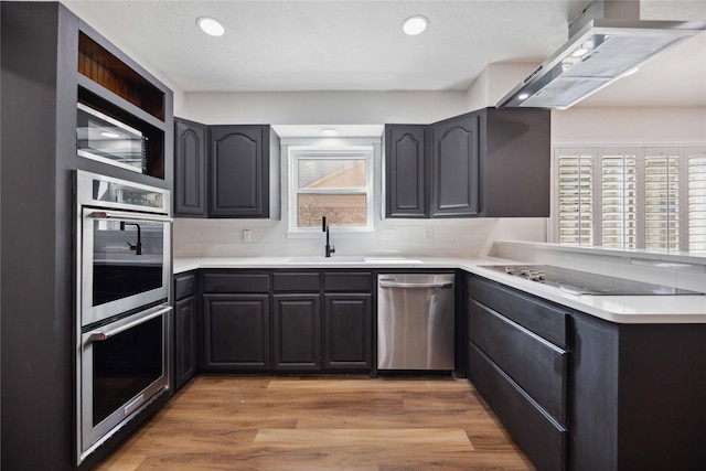 kitchen with stainless steel appliances, a sink, light wood-style floors, backsplash, and exhaust hood