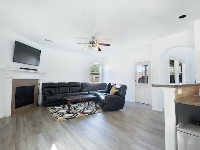 living area featuring visible vents, ceiling fan, a tiled fireplace, arched walkways, and light wood-style floors