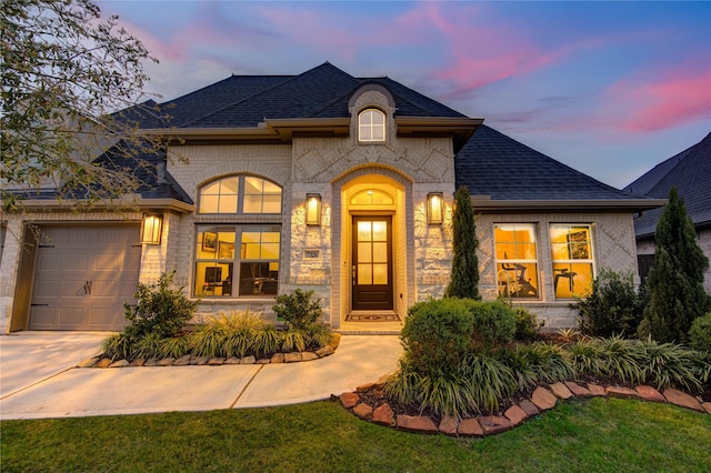 french country home with roof with shingles, an attached garage, brick siding, and concrete driveway