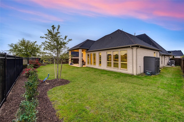 rear view of house with central AC unit, a fenced backyard, brick siding, a lawn, and a shingled roof