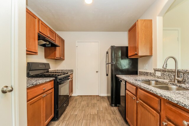 kitchen with a sink, light wood finished floors, light stone countertops, under cabinet range hood, and black appliances