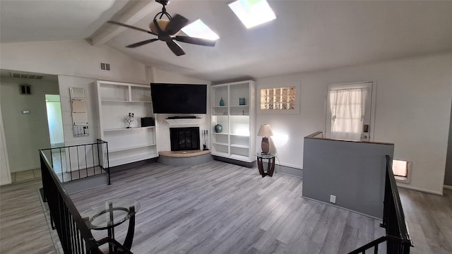 living room featuring wood finished floors, a glass covered fireplace, visible vents, a ceiling fan, and lofted ceiling with skylight