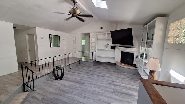 living area featuring wood finished floors, ceiling fan, a fireplace with raised hearth, and vaulted ceiling with skylight
