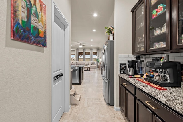 kitchen with stainless steel appliances, backsplash, light stone countertops, dark brown cabinets, and glass insert cabinets