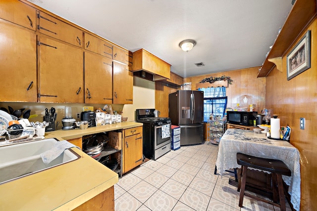 kitchen with light tile patterned floors, appliances with stainless steel finishes, wood walls, light countertops, and brown cabinetry