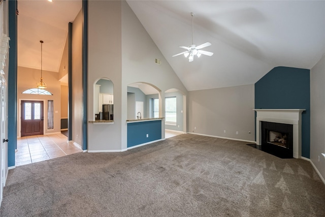 unfurnished living room featuring baseboards, a ceiling fan, a fireplace with flush hearth, high vaulted ceiling, and light colored carpet