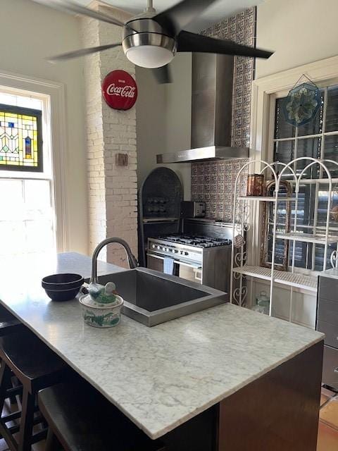 kitchen with a sink, wall chimney range hood, stainless steel stove, a ceiling fan, and decorative backsplash