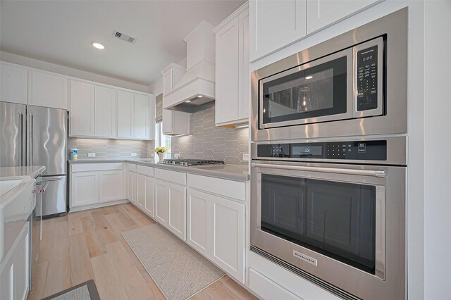2611 Oakland Park Dr, Conroe, TX 77385 - kitchen with appliances with stainless steel finishes, light wood finished floors, custom range hood, tasteful backsplash, and white cabinets
