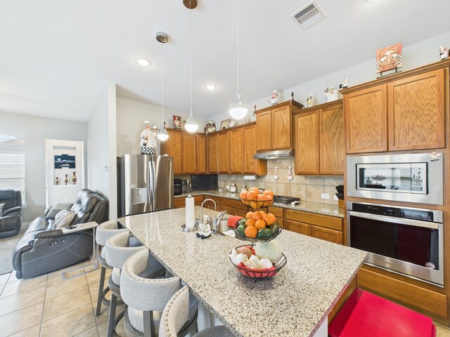 kitchen featuring decorative backsplash, a kitchen breakfast bar, stainless steel appliances, under cabinet range hood, and brown cabinetry