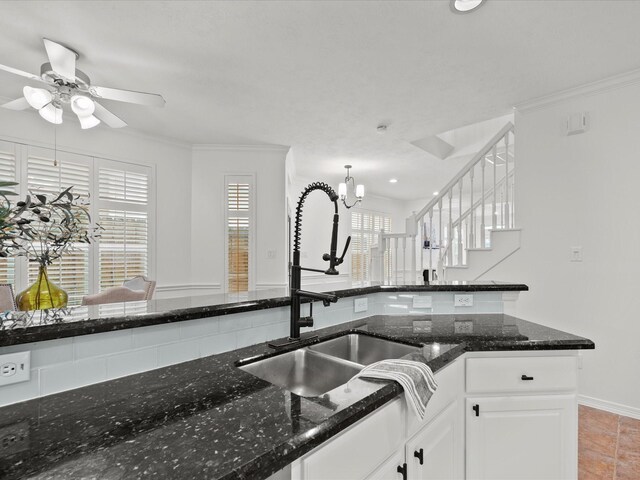 kitchen with a sink, white cabinetry, dark stone countertops, ceiling fan with notable chandelier, and ornamental molding