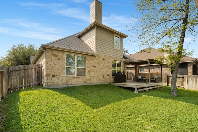 back of property with stone siding, a deck, a fenced backyard, a shingled roof, and a yard