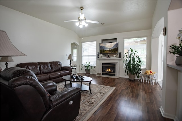 living room with ceiling fan, dark wood-style flooring, a warm lit fireplace, arched walkways, and baseboards