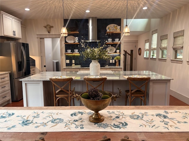 kitchen with wall chimney range hood, light stone counters, open shelves, a spacious island, and stainless steel refrigerator with ice dispenser