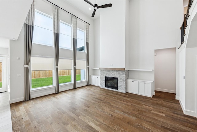 unfurnished living room with a towering ceiling, wood finished floors, a stone fireplace, a ceiling fan, and baseboards