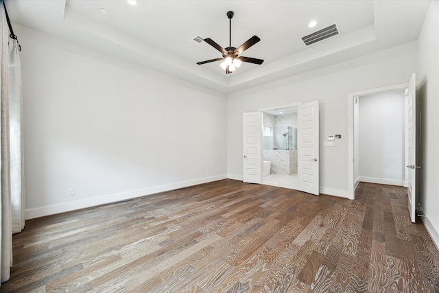 unfurnished bedroom featuring wood finished floors, a tray ceiling, baseboards, ensuite bath, and visible vents