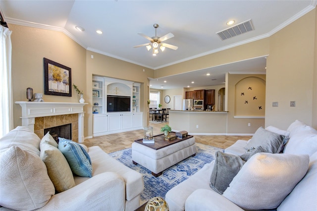 living area with built in features, a tiled fireplace, crown molding, visible vents, and ceiling fan