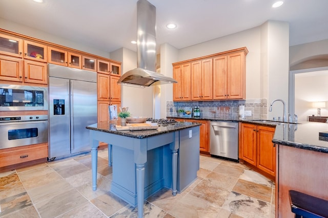 kitchen with built in appliances, island range hood, a sink, dark stone counters, and a breakfast bar