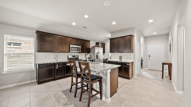 kitchen featuring dark brown cabinetry, light stone counters, stainless steel appliances, a center island with sink, and backsplash