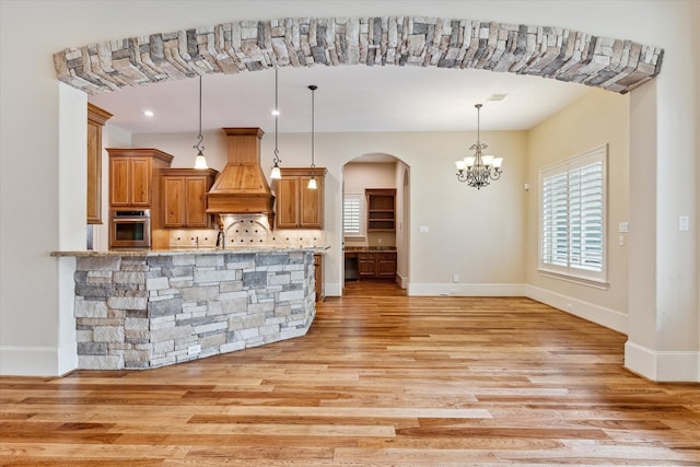 kitchen with light wood finished floors, brown cabinets, a notable chandelier, oven, and premium range hood