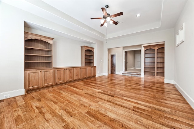 unfurnished living room with a tray ceiling, ceiling fan, baseboards, and light wood-style floors