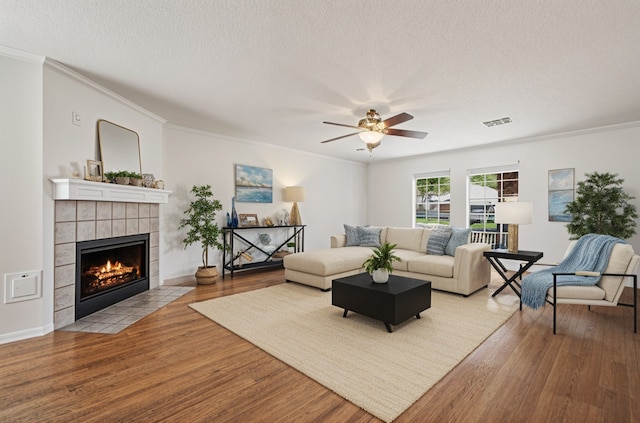 living room featuring a tile fireplace, ceiling fan, crown molding, visible vents, and wood finished floors