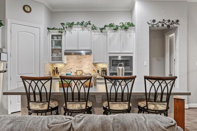137 Gray Vervain Ct, Montgomery, TX 77316 - kitchen with white cabinets, stainless steel microwave, backsplash, a breakfast bar, and under cabinet range hood