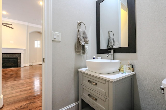 half bathroom with vanity, baseboards, a tiled fireplace, a ceiling fan, and wood finished floors
