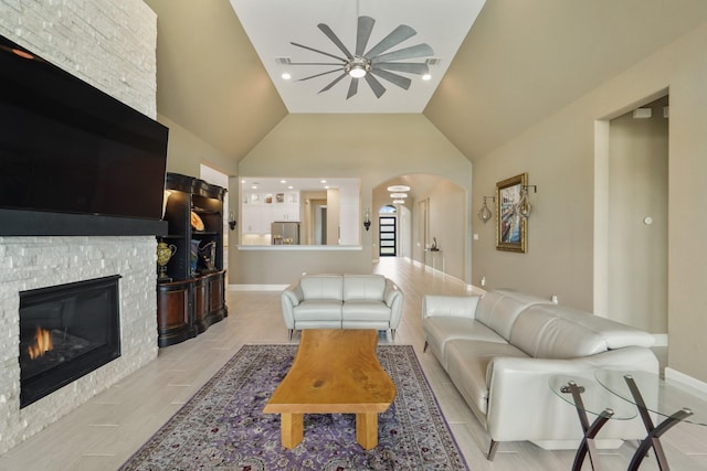 living room featuring vaulted ceiling, a ceiling fan, a stone fireplace, arched walkways, and baseboards