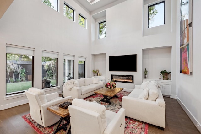 living room featuring dark wood-style flooring, baseboards, a glass covered fireplace, and a towering ceiling
