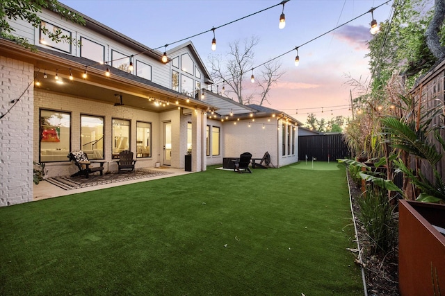 back of house at dusk with a patio, a lawn, fence, and brick siding