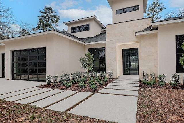 entrance to property with stucco siding, stone siding, a garage, and roof with shingles