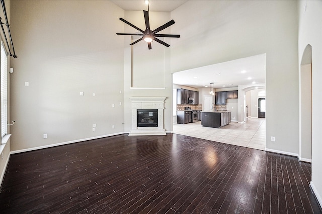 unfurnished living room featuring light wood-style flooring, arched walkways, baseboards, a high ceiling, and ceiling fan