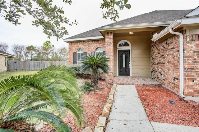 doorway to property featuring brick siding, fence, and a shingled roof