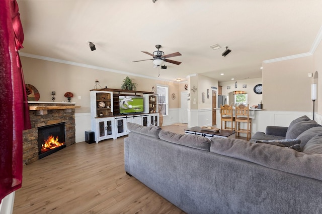 living room featuring a stone fireplace, ceiling fan, crown molding, wainscoting, and wood finished floors