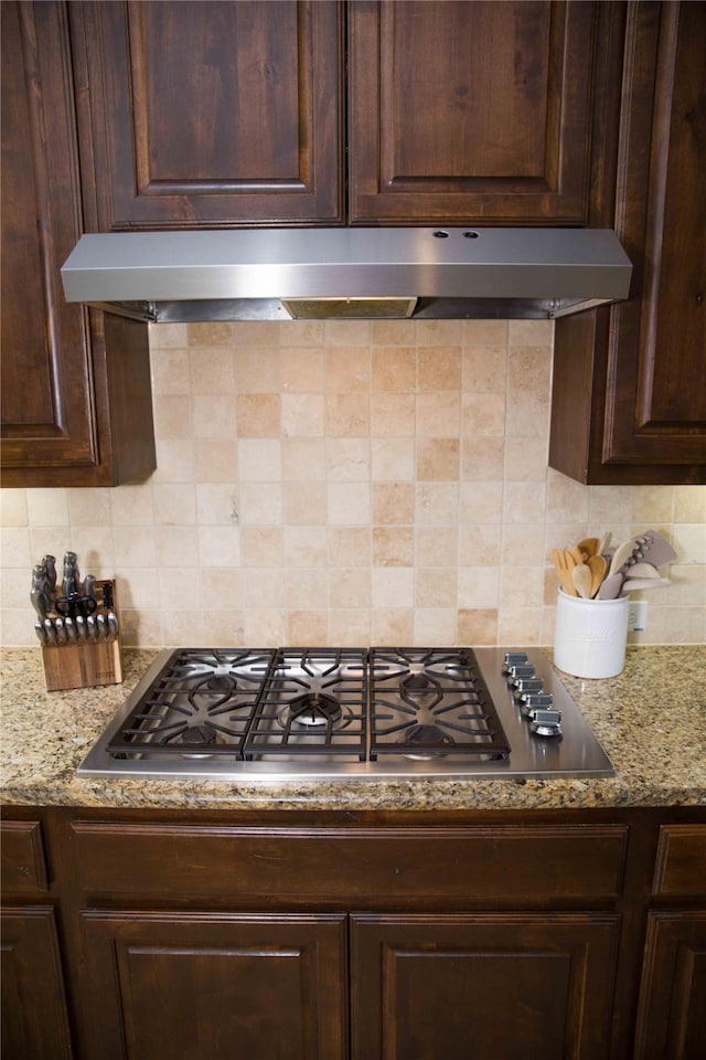 kitchen with dark brown cabinets, light stone counters, decorative backsplash, stainless steel gas stovetop, and under cabinet range hood