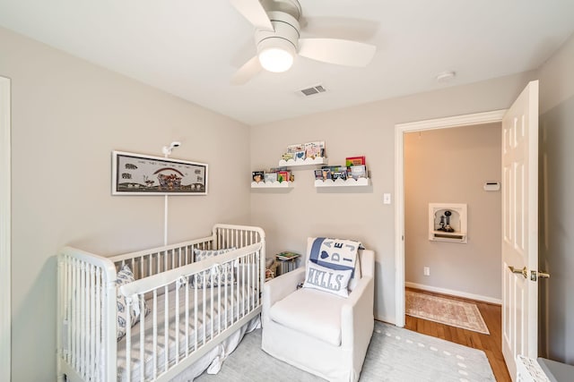 bedroom featuring ceiling fan, visible vents, wood finished floors, a crib, and baseboards