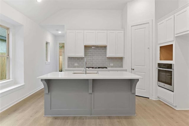 kitchen with an island with sink, stainless steel oven, white cabinetry, a sink, and light countertops