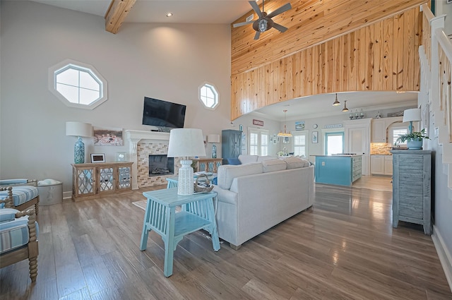 living area featuring ceiling fan, a high ceiling, wood finished floors, a tiled fireplace, and beam ceiling