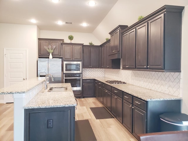 kitchen with a kitchen island with sink, backsplash, appliances with stainless steel finishes, light wood-type flooring, and a sink