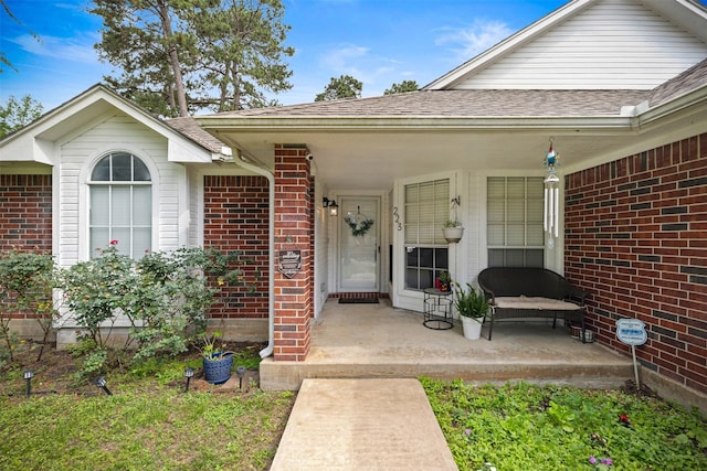 doorway to property with a shingled roof, a porch, and brick siding