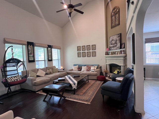 living room featuring a ceiling fan, arched walkways, baseboards, high vaulted ceiling, and a glass covered fireplace