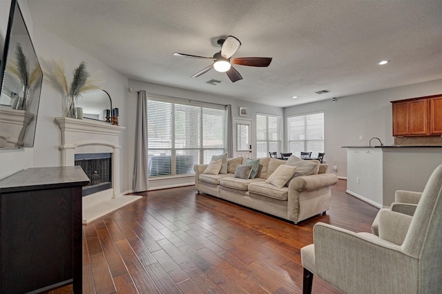 living area featuring a ceiling fan, dark wood-type flooring, a fireplace with raised hearth, baseboards, and a textured ceiling